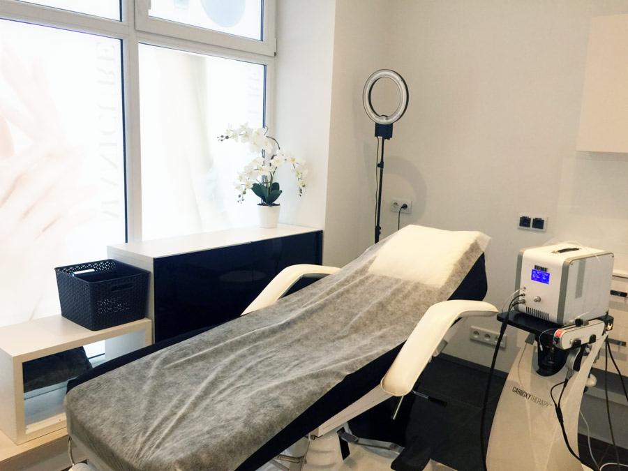Jasne wnętrze gabinetu z łóżkiem zabiegowym przykrytym jednorazowym prześcieradłem, obok widoczny sprzęt do karboksyterapii, na ścianie okrągła lampa doświetlająca, a w tle okno z dekoracją.