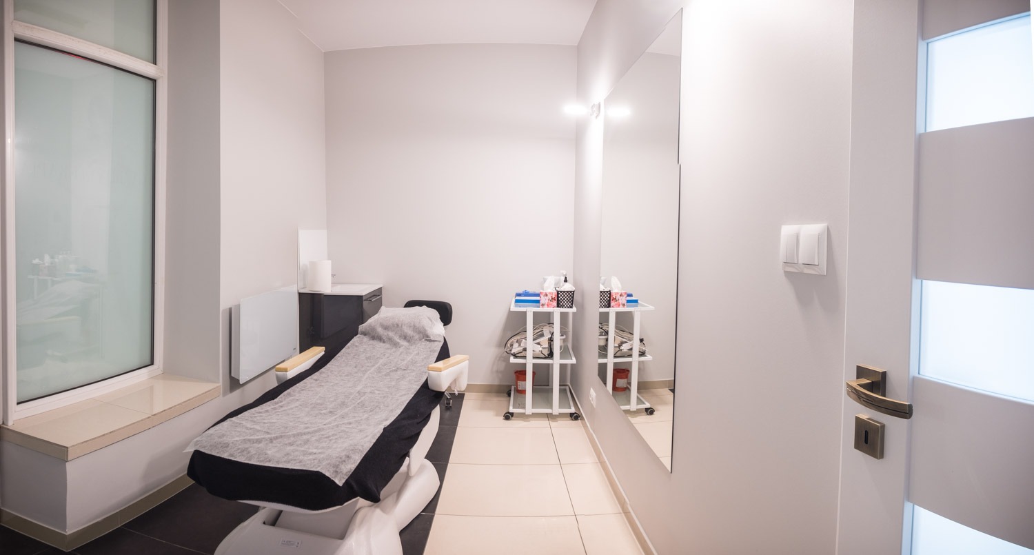 Jasne wnętrze gabinetu z kozetką przykrytą szarym prześcieradłem, w tle wózek z akcesoriami i duże lustro.