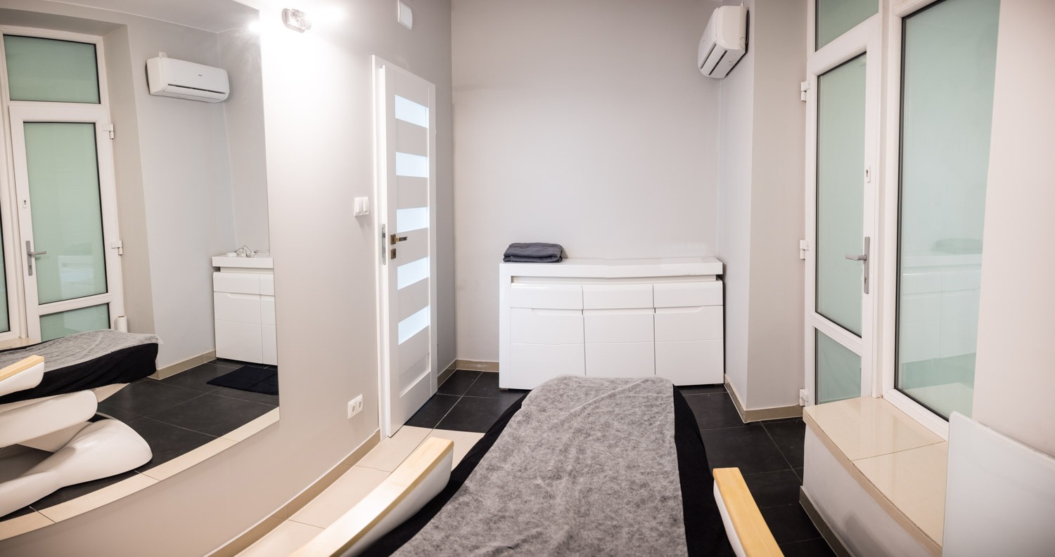 Jasne wnętrze gabinetu z łóżkiem do zabiegów, białymi meblami i dużym lustrem, odbijającym dodatkowe stanowisko.
