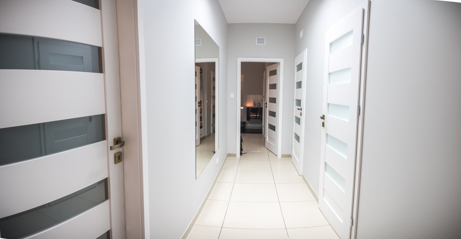 Wnętrze korytarza w jasnych szarościach z białymi drzwiami z przeszkleniami i dużym lustrem na ścianie, widok w głąb pomieszczenia.