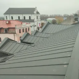 Nowy, szary dach z blachy na tle zabudowy miejskiej z kościołem w tle, widoczne okna dachowe.