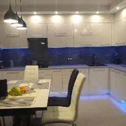 Nowoczesna kuchnia z białymi, lakierowanymi szafkami, podświetleniem LED pod szafkami wiszącymi i dolnymi, szarym blatem i marmurowym panelem nad blatem, widok na stół z laptopem i owocami.