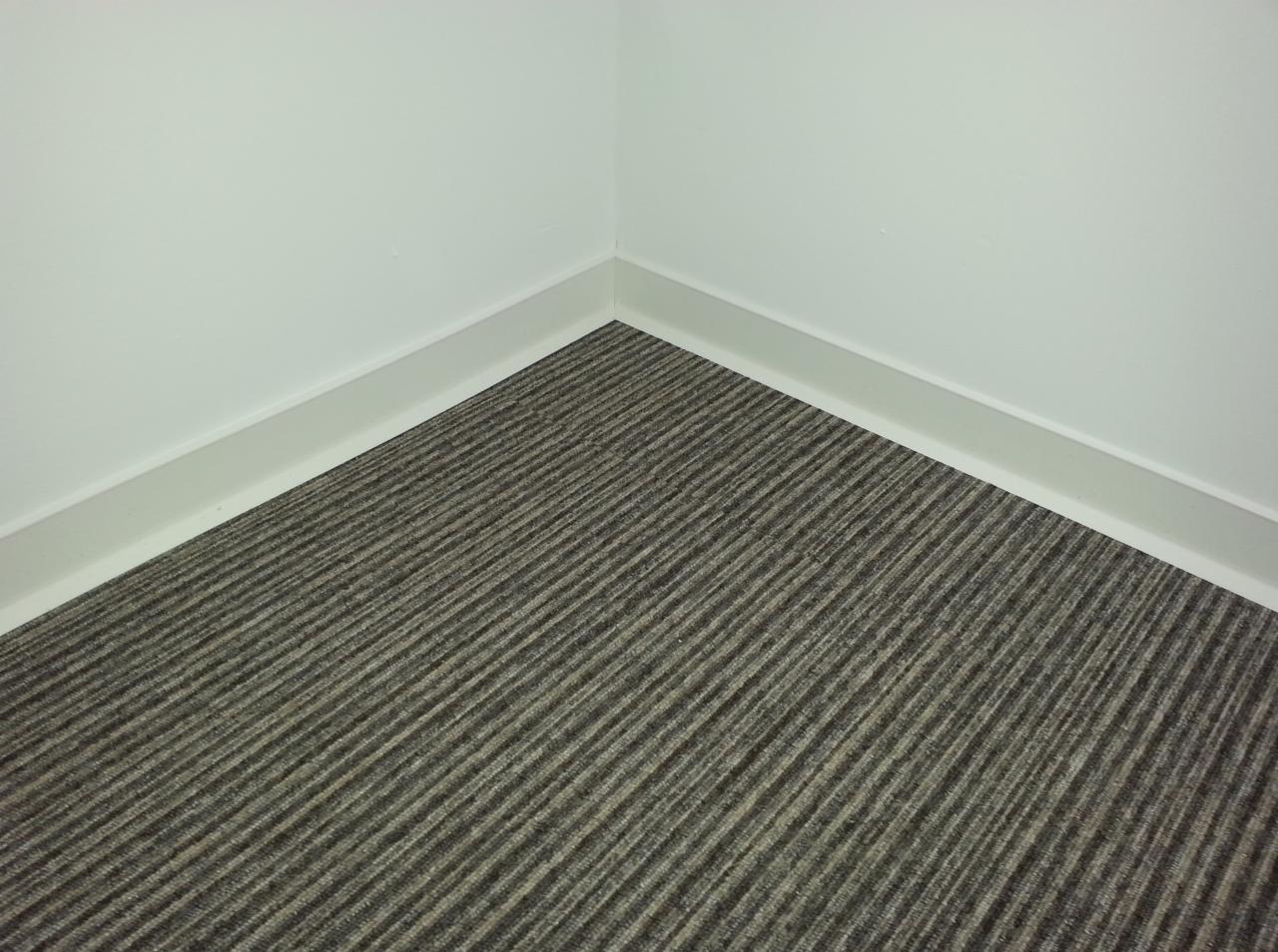 Wykładzina dywanowa w odcieniach szarości z subtelnym wzorem prążków, ułożona w rogu pomieszczenia w Wężyczynie, z widocznymi białymi listwami przypodłogowymi.