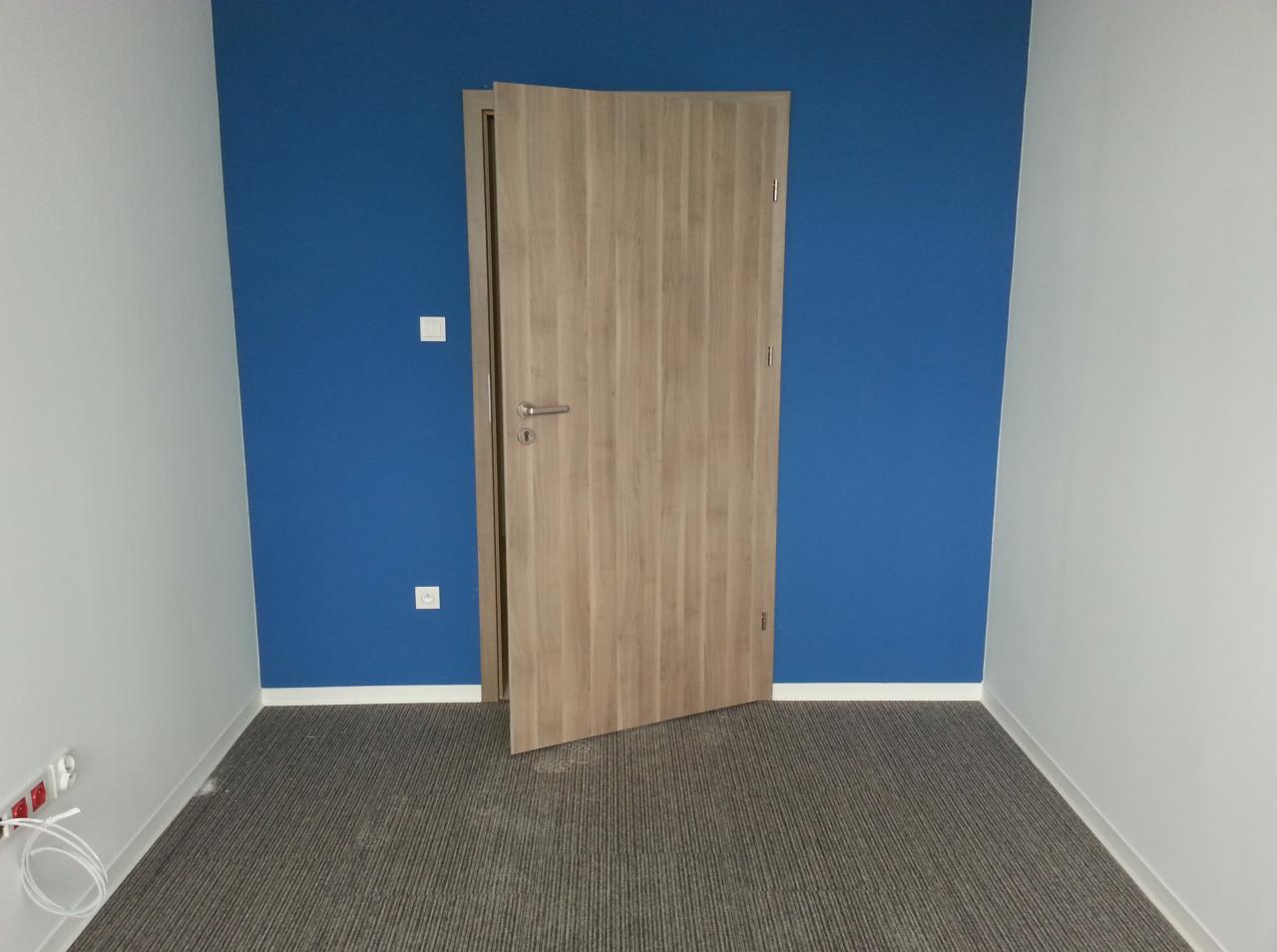 Pokój z szarą wykładziną dywanową w paski, niebieską ścianą i częściowo otwartymi drzwiami z drewnianym fornirem.