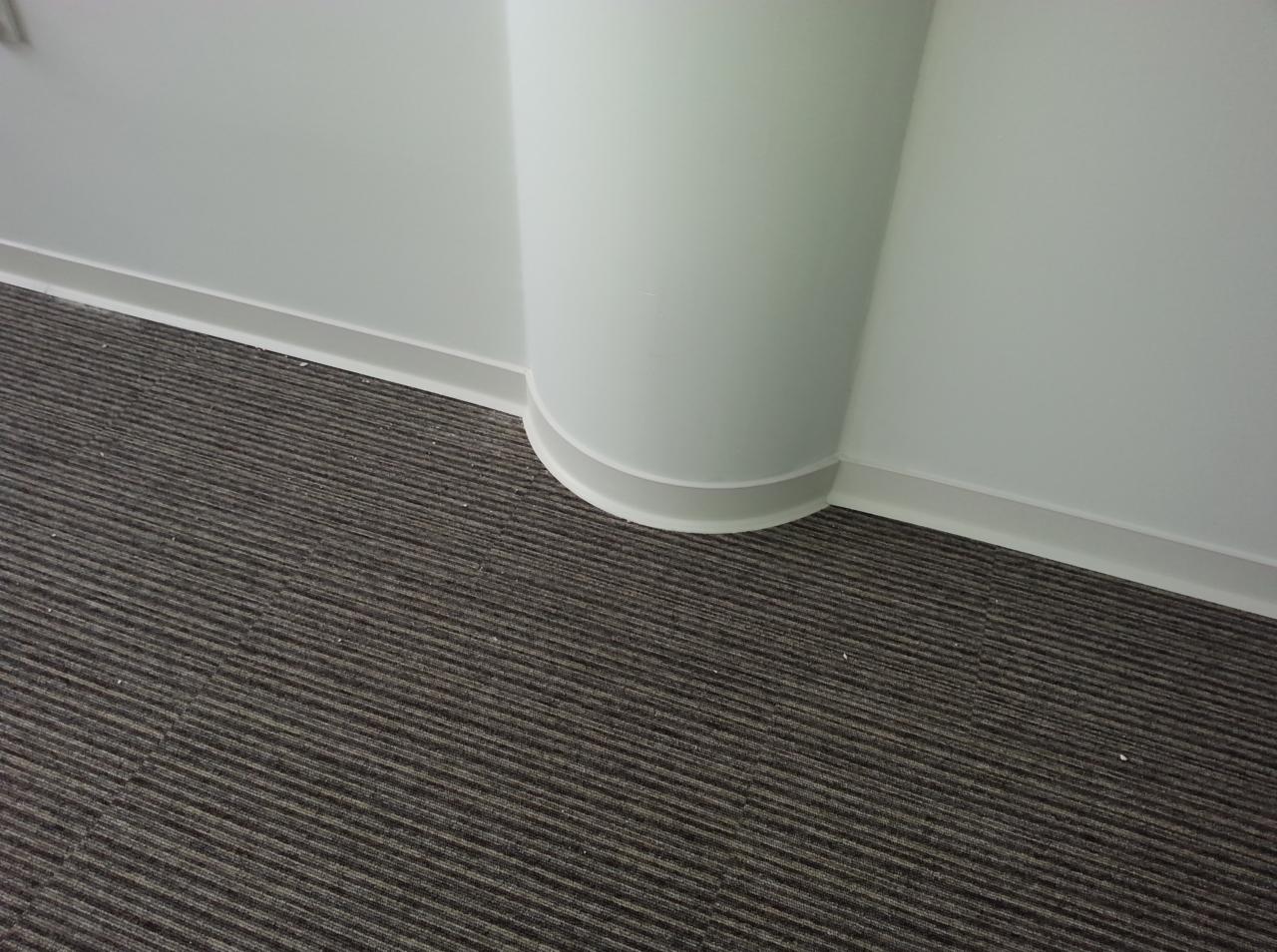 Szara wykładzina dywanowa w paski przy białej kolumnie i listwie przypodłogowej.