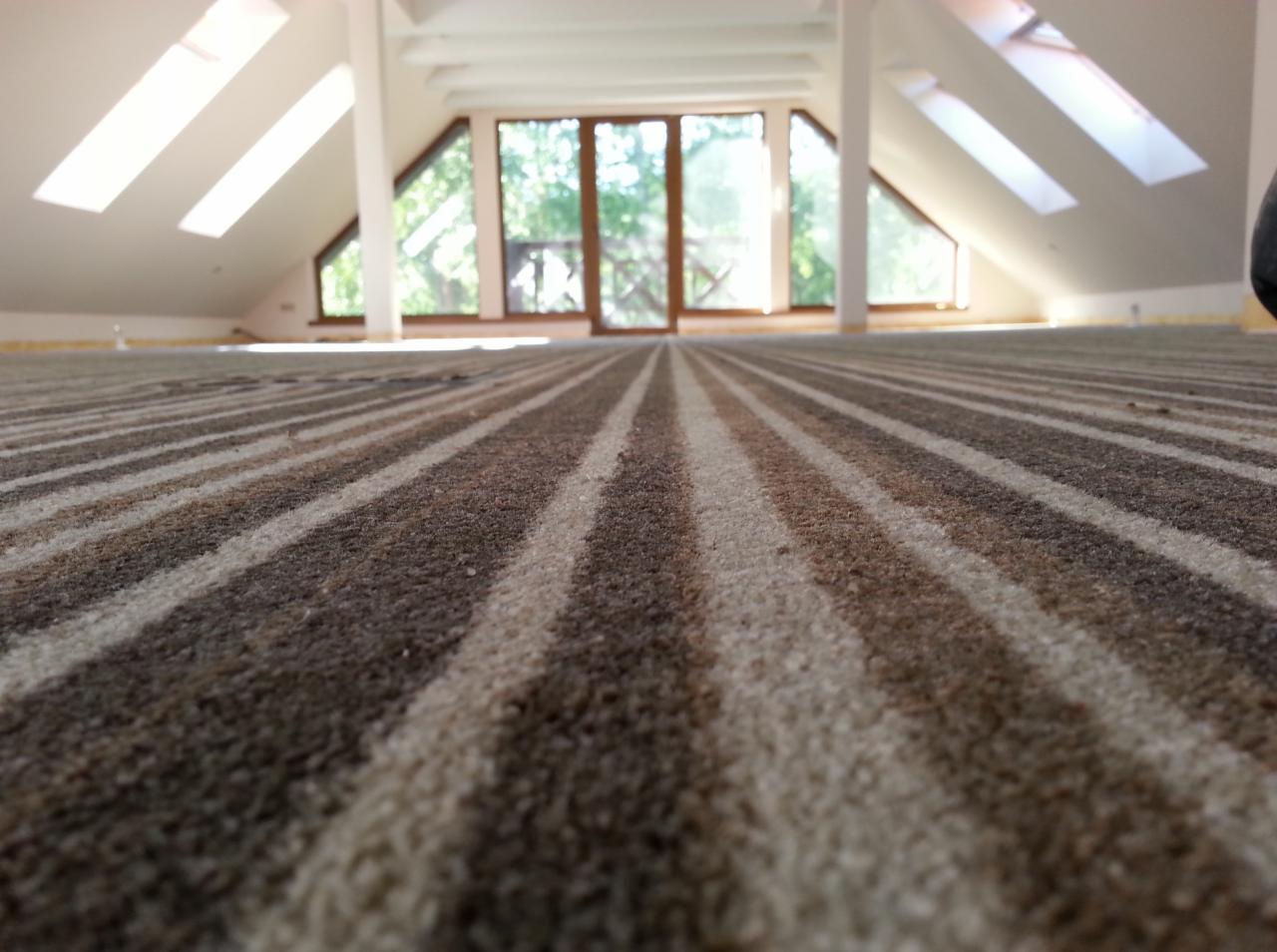 Pasy brązowo-beżowej wykładziny dywanowej w jasnym pokoju na poddaszu z oknami dachowymi i dużym oknem na wprost.