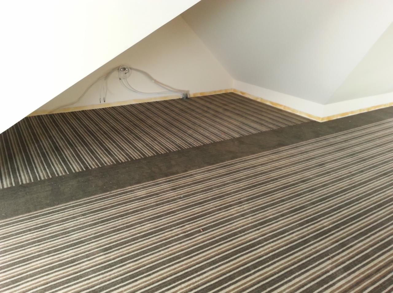 Wnętrze poddasza z nową wykładziną w paski, widoczne kable przy listwie przypodłogowej, żółta listwa wykończeniowa.