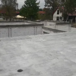 Wylana płyta fundamentowa z wystającymi prętami zbrojeniowymi, bloczki betonowe tworzące ściany fundamentowe, widok na dom w tle.