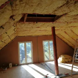 Poddasze w trakcie izolacji termicznej wełną mineralną, widoczne krokwie, okna dachowe i fragment rusztowania. W tle widok na zieleń za oknem.