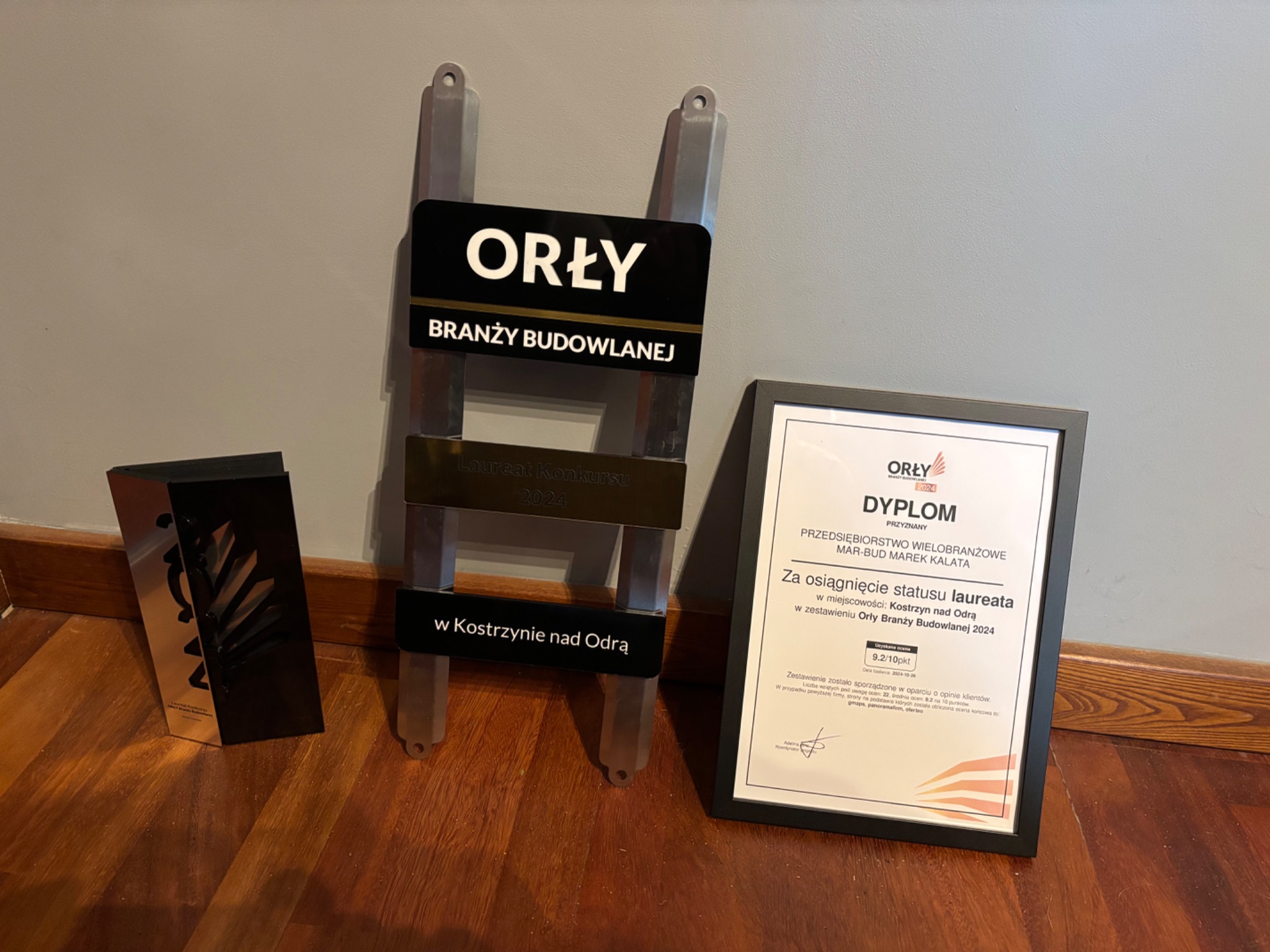 Nagrody Orły Branży Budowlanej 2024 dla firmy MAR-BUD Marek Kalata, w tym statuetka w kształcie drabiny, dyplom w czarnej ramce i druga statuetka na drewnianej podłodze.