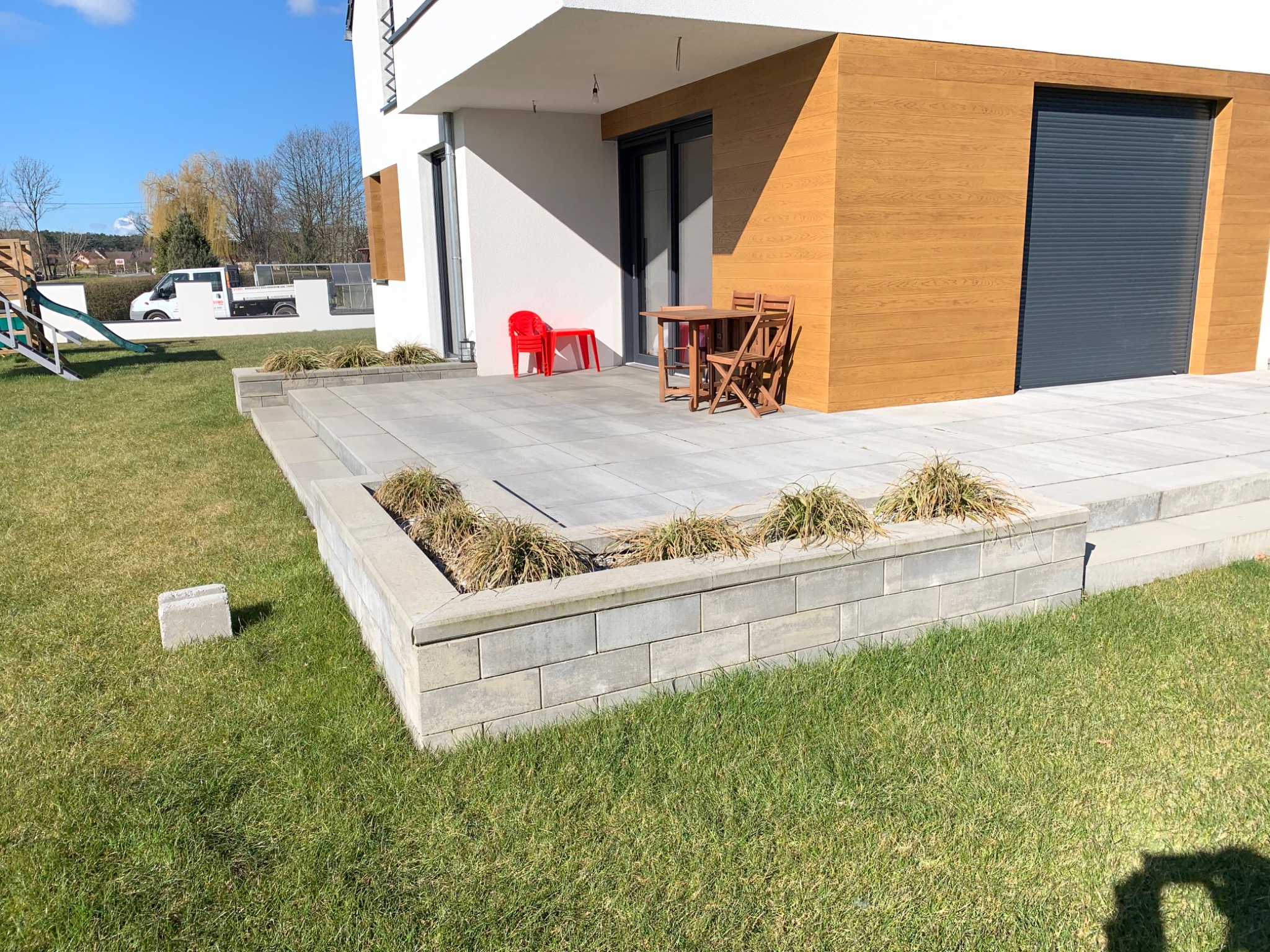 Nowoczesny taras z szarej kostki brukowej, otoczony rabatą z suchymi trawami, z widocznym fragmentem budynku z drewnianą elewacją i szarą bramą garażową oraz czerwonym krzesłem i drewnianym...