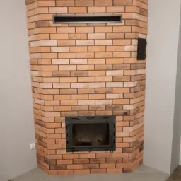 Obudowa kominka z cegły klinkierowej w odcieniach brązu i pomarańczy, z czarną wkładką kominkową i kratką wentylacyjną u góry, na tle szarej ściany i podłogi z płytek.