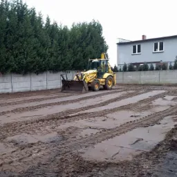 Żółta koparko-ładowarka Komatsu wyrównuje błotnisty teren z widocznymi śladami opon, w tle betonowy płot i budynek mieszkalny.