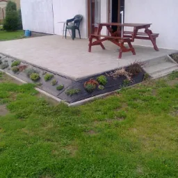 Wyłożony kostką brukową taras z niskim rabatem obsadzonym roślinami, z drewnianym stołem piknikowym i krzesłem z tworzywa sztucznego, widok na dom z białymi ścianami.