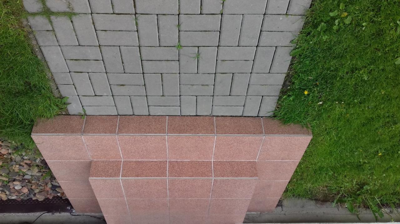 Widok z góry na ułożoną kostkę brukową w szarym odcieniu, graniczącą z trawnikiem, oraz fragment schodów wyłożonych płytkami w odcieniach brązu i beżu. W tle widoczny fragment kamieni ozdobnych.