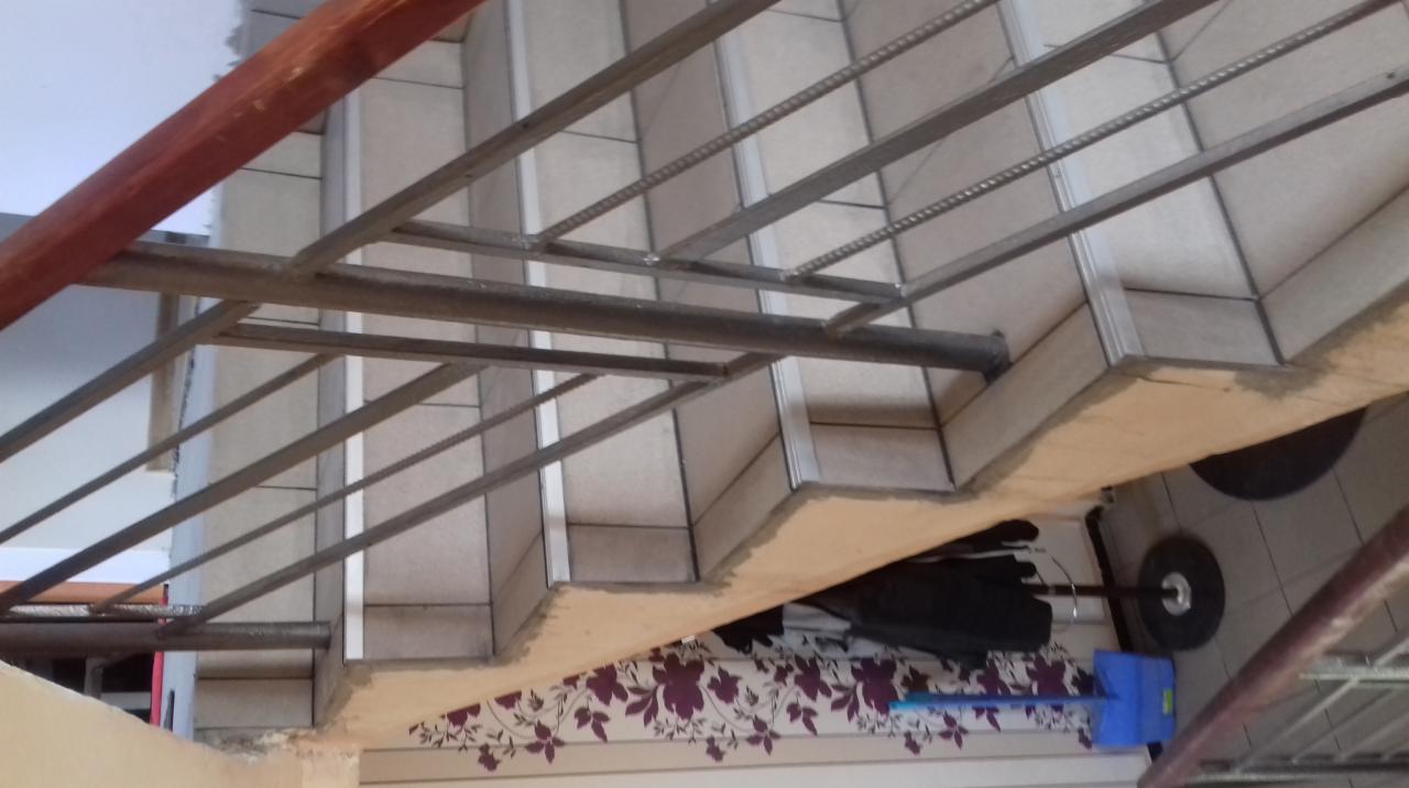 Metalowa konstrukcja schodów z widocznymi elementami spawania, płytki ceramiczne na stopniach, fragment ściany z wzorem kwiatowym w tle, ujęcie z dołu.