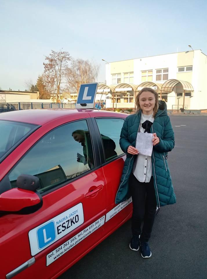 Uśmiechnięta dziewczyna trzymająca dokument, stojąca obok czerwonego samochodu z literą 'L' na dachu i logo firmy na drzwiach.