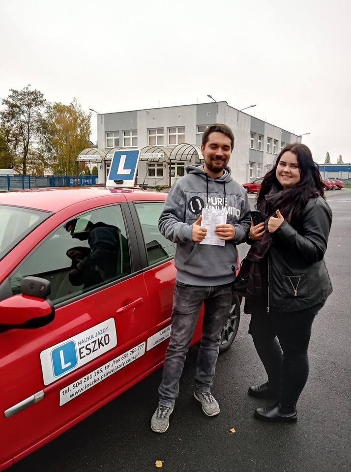 Uśmiechnięty mężczyzna z dokumentem i kobieta z telefonem, oboje pokazują kciuk w górę, stojący obok czerwonego samochodu z naklejką informacyjną firmy oferującej kursy nauki jazdy.