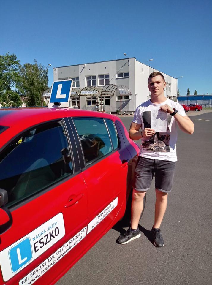 Młody mężczyzna trzymający dokument, stojący obok czerwonego samochodu szkoły jazdy z literą 'L' na dachu i naklejką 'Nauka Jazdy Leszko', na tle budynku szkoły.