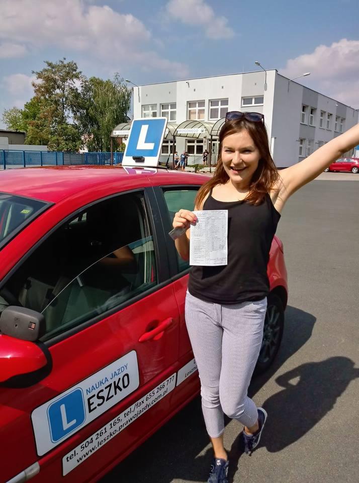 Uśmiechnięta kobieta z uniesioną ręką trzyma dokument przed czerwonym samochodem z literą 'L' na dachu, oznaczającym naukę jazdy, na tle budynku.