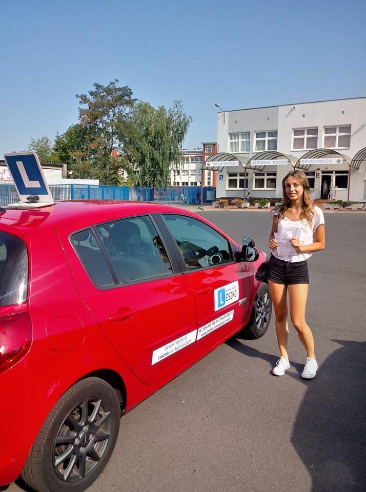 Uśmiechnięta młoda kobieta trzymająca dokumenty stoi obok czerwonego samochodu z literą 'L' na dachu, na tle budynku Ośrodka Ruchu Drogowego.
