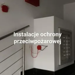 Instalacja przeciwpożarowa w klatce schodowej: centrala alarmowa, czujniki pożarowe, metalowe schody z czarnymi stopniami.