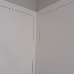 USŁUGI BUDOWLANE MONTERS ŁUKASZ SZYBAJŁO - Dwie białe, minimalistyczne drzwi wewnętrzne z czarnymi klamkami, ustawione pod kątem w rogu pomieszczenia, widoczne z bliska.