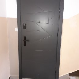 USŁUGI BUDOWLANE MONTERS ŁUKASZ SZYBAJŁO - Grafitowe drzwi wejściowe z geometrycznym wzorem, czarną klamką i wizjerem, zamontowane w klatce schodowej z beżowymi ścianami.