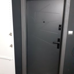 USŁUGI BUDOWLANE MONTERS ŁUKASZ SZYBAJŁO - Szare, nowoczesne drzwi wejściowe z geometrycznym wzorem, czarną klamką i wizjerem, zamontowane w ciemnoszarym murze, obok biała szafa z widocznymi zawiasami.