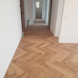 Ułożona podłoga z paneli w jodełkę, widok na korytarz z białymi ścianami i butelką wody przy ścianie.