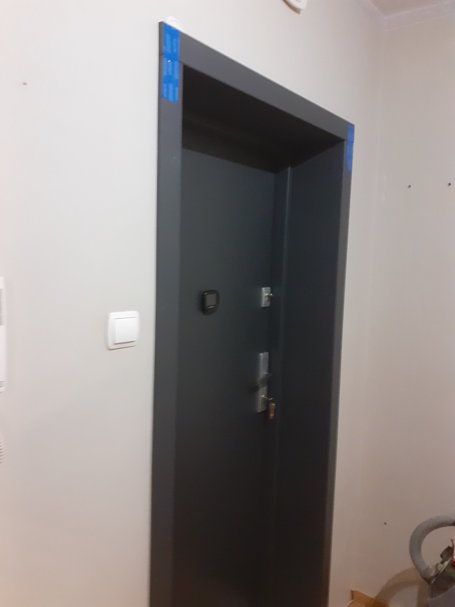 Ciemnoszare drzwi wejściowe z zamontowanym wizjerem i zabezpieczeniami, zabezpieczone niebieską taśmą malarską na ościeżnicy podczas montażu w jasnym wnętrzu.