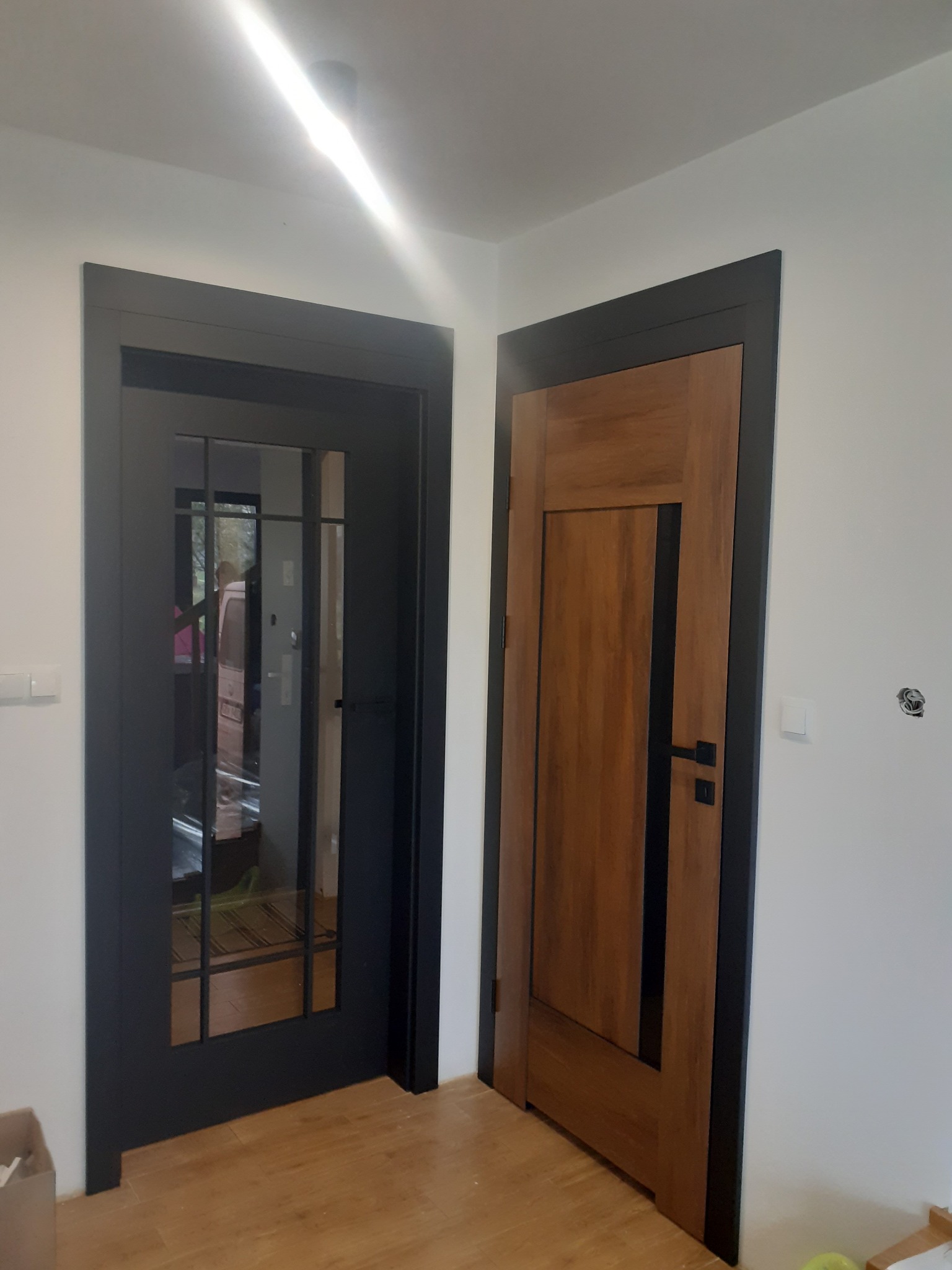 Dwa różne modele drzwi wewnętrznych z czarnymi ościeżnicami: przeszklone drzwi z kratką i drewniane drzwi z pionowym, czarnym paskiem, zamontowane w narożniku pomieszczenia z jasnymi ścianami...