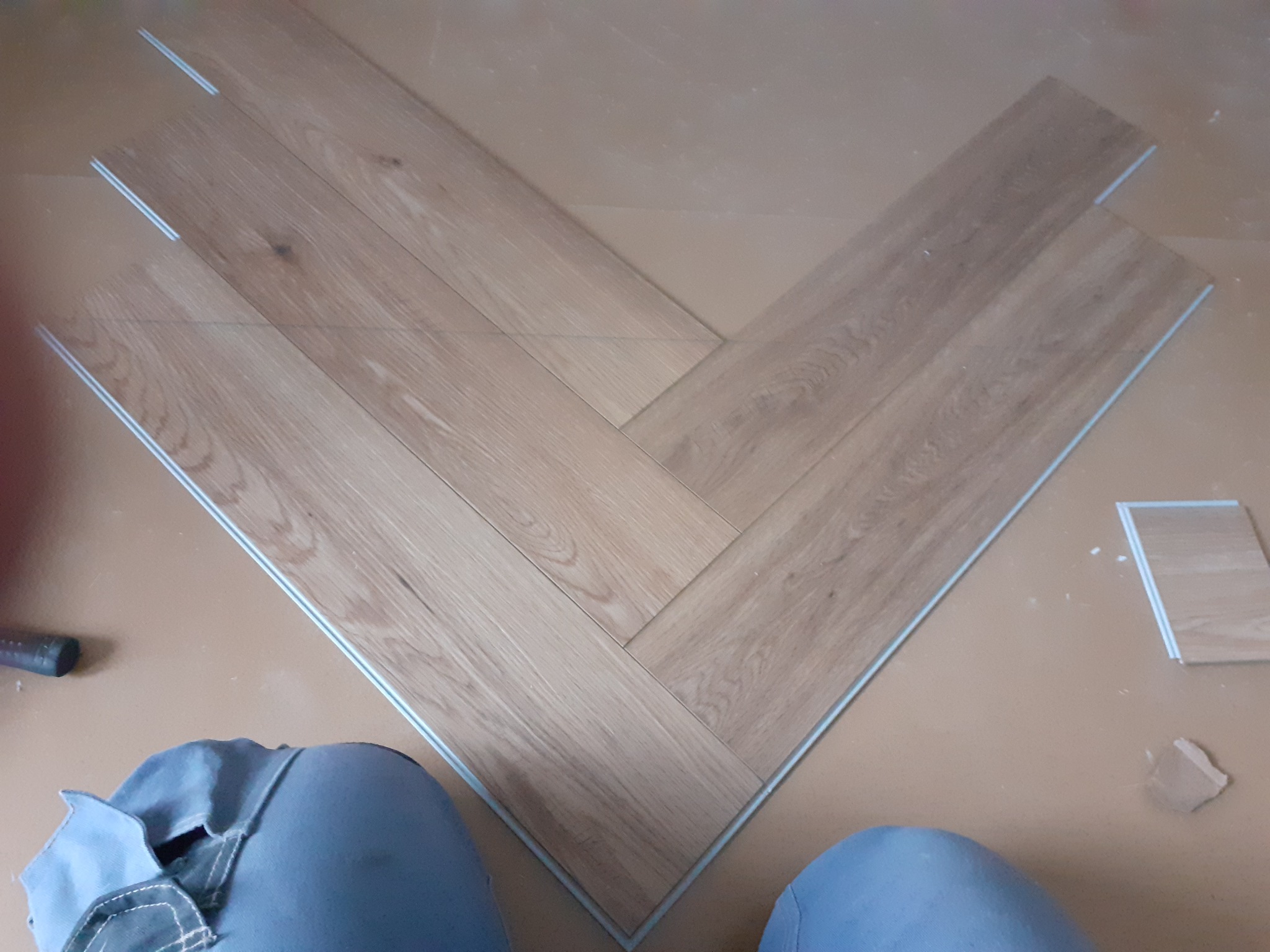 Fragment podłogi z jasnych paneli ułożonych we wzór jodełki, widoczne białe krawędzie łączeń, w tle fragment ubrania osoby montującej i narzędzie.