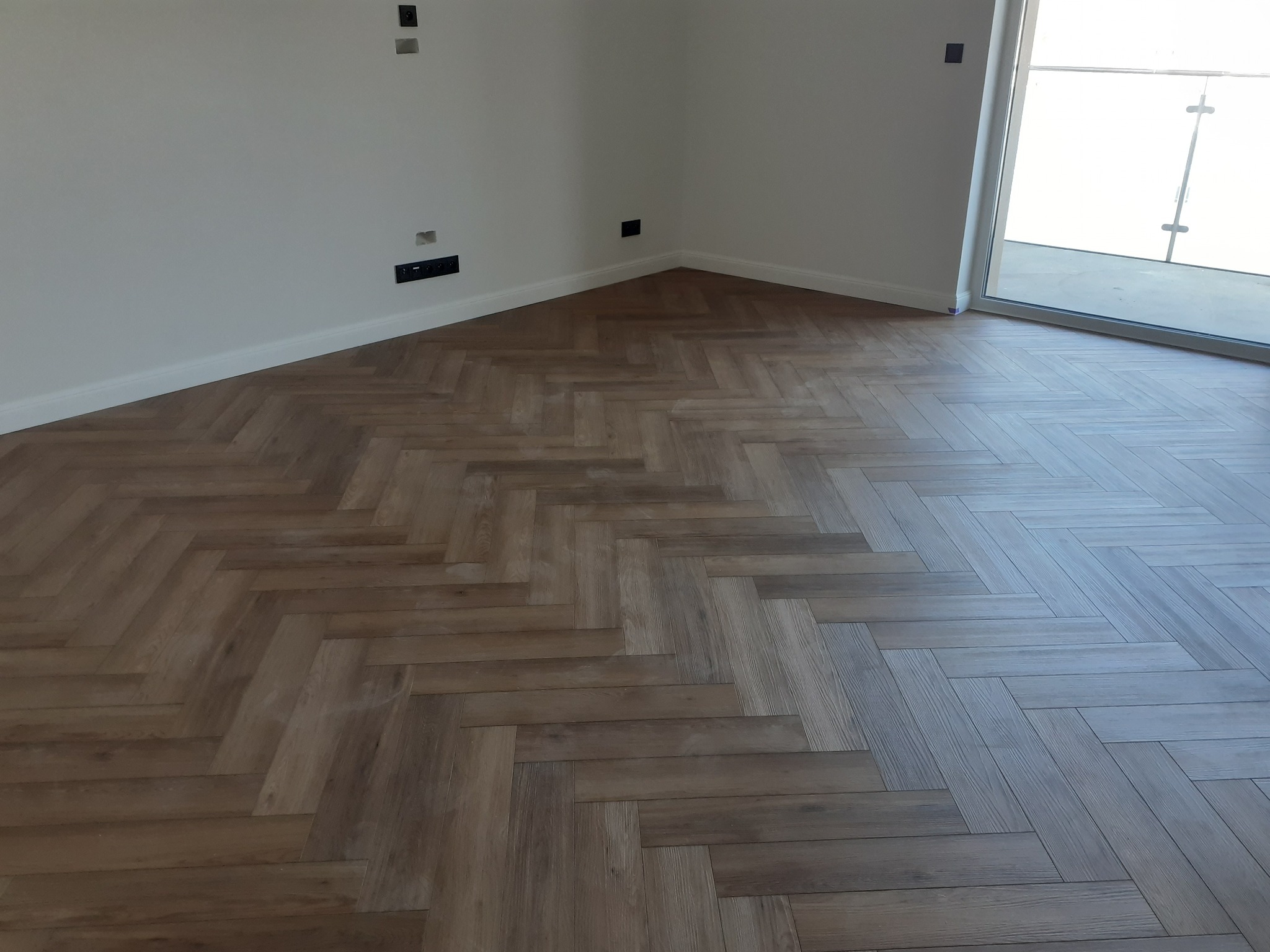 Ukończona podłoga z paneli ułożonych w jodełkę, widoczne gniazdka elektryczne na białej ścianie i fragment drzwi balkonowych.