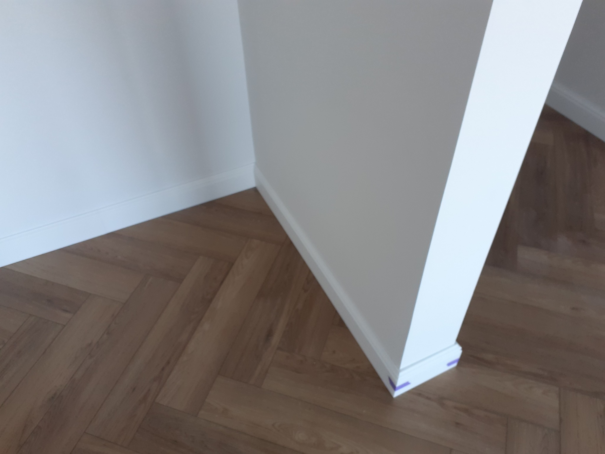 Ukończone układanie parkietu w jodełkę francuską, widoczne listwy przypodłogowe i fragment ściany z narożnikiem, zabezpieczone niebieską taśmą malarską.