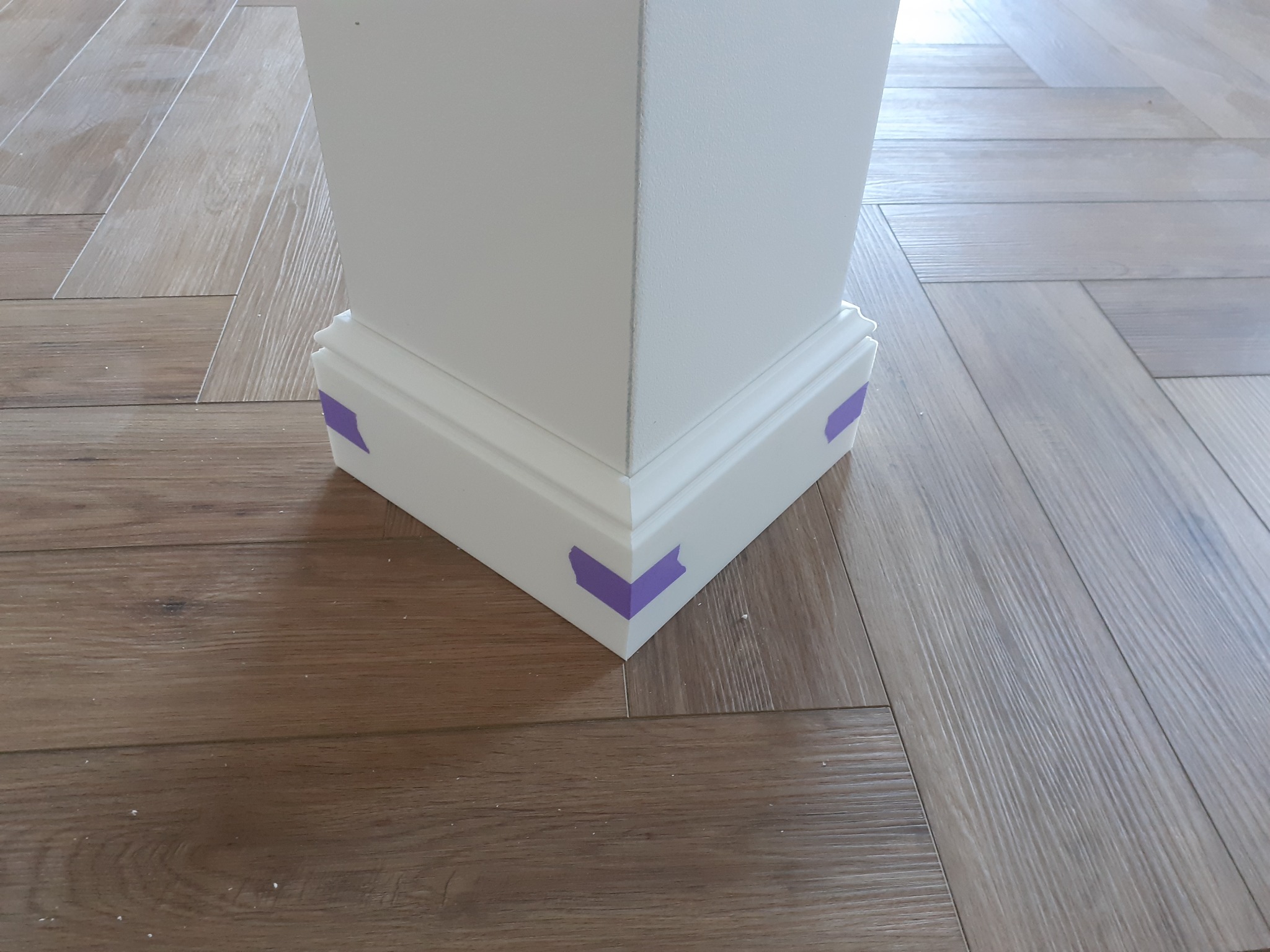 Fragment podłogi z paneli ułożonych w jodełkę przy białej kolumnie z listwą przypodłogową zabezpieczoną fioletową taśmą malarską podczas remontu w Kwidzynie.