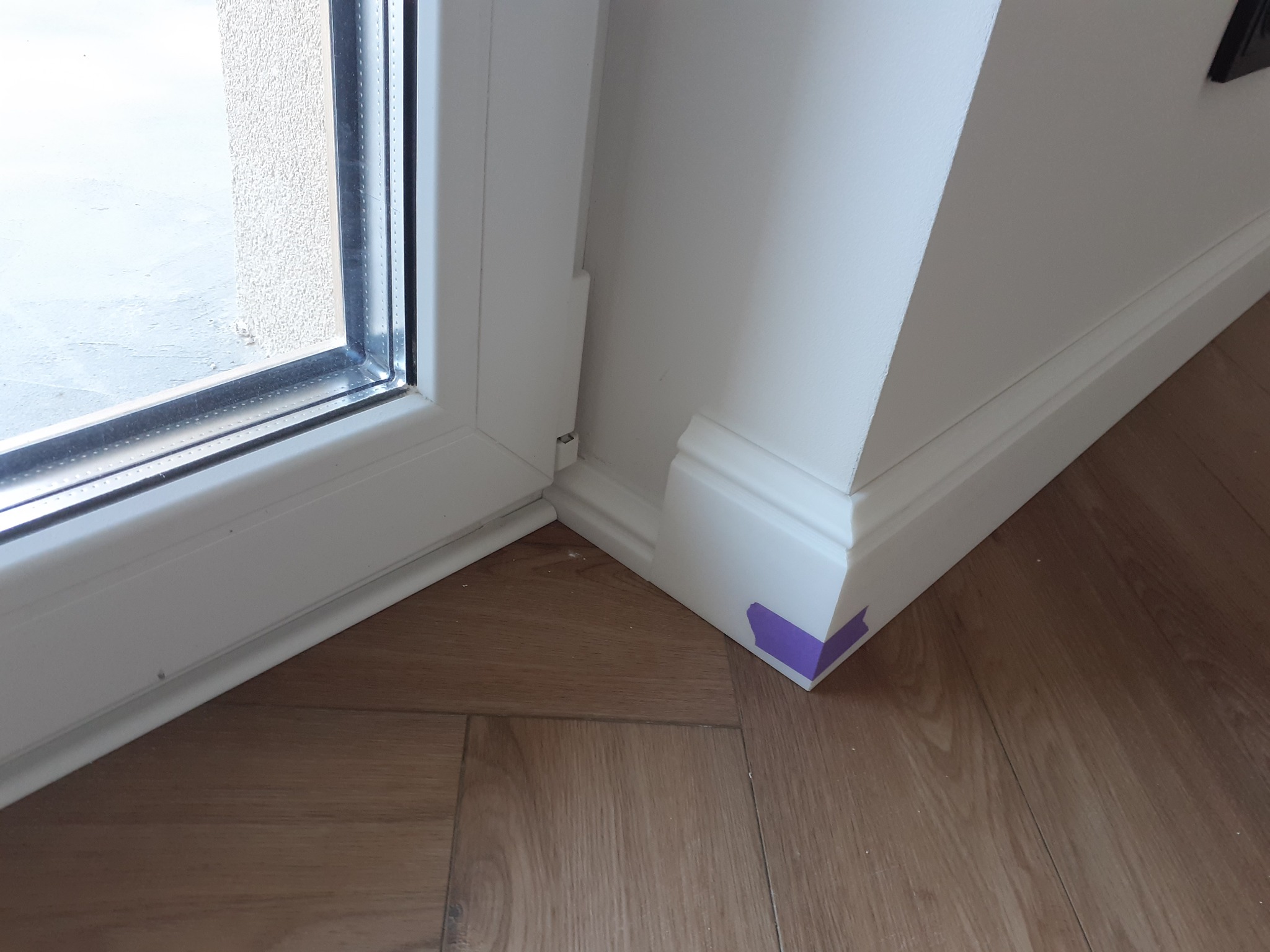 Fragment pomieszczenia z drewnopodobną podłogą, białą listwą przypodłogową zabezpieczoną fioletową taśmą malarską w rogu, oraz fragmentem białej ramy okiennej.