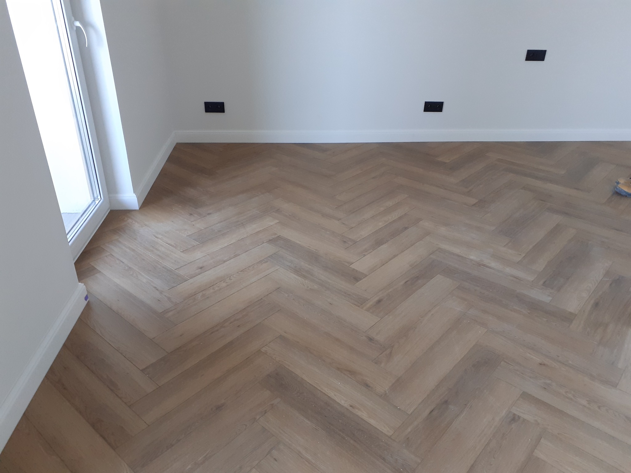 Ułożona podłoga z paneli w jodełkę, widoczne czarne gniazdka elektryczne na białej ścianie i białe listwy przypodłogowe.