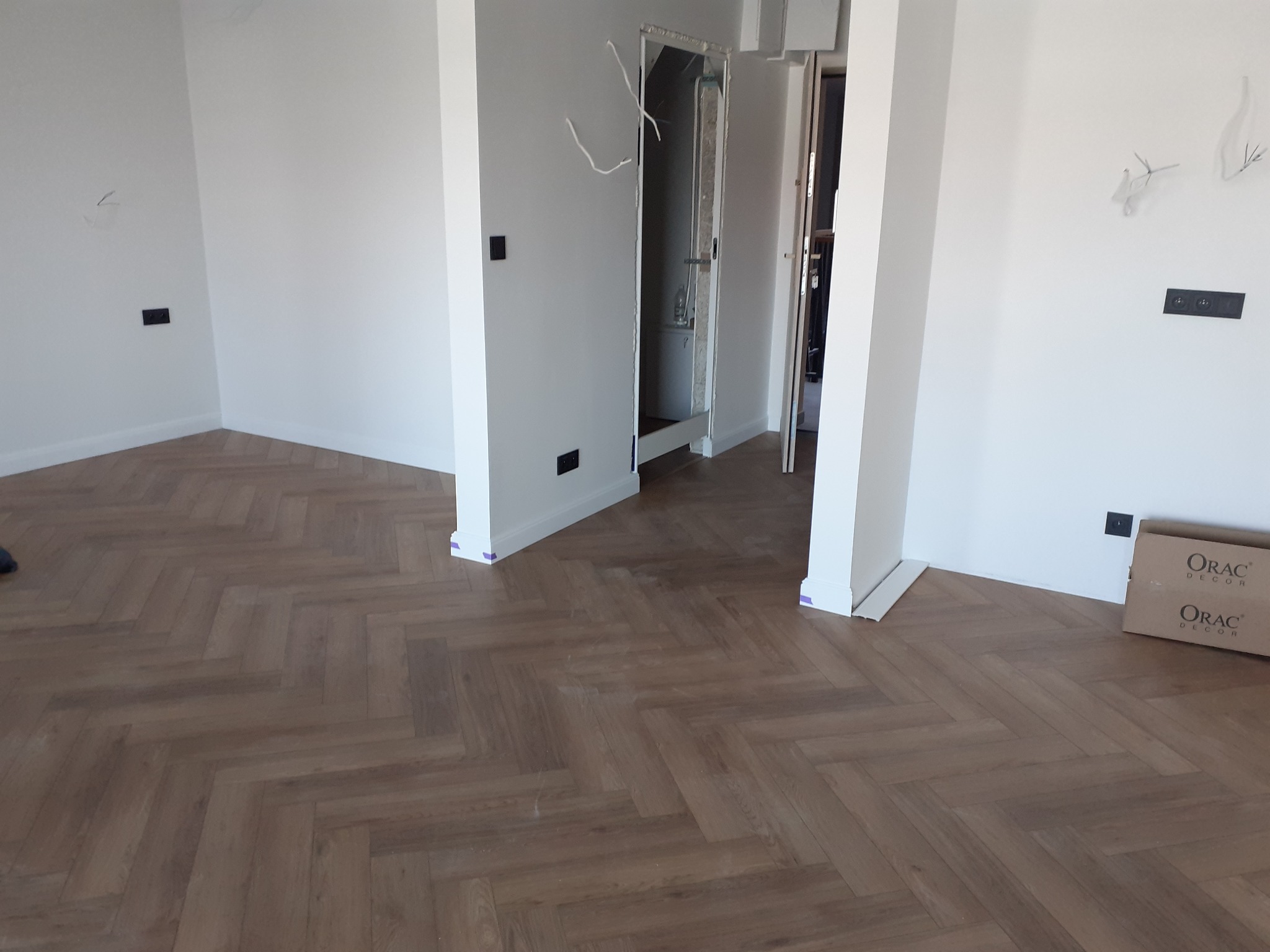 Ułożona podłoga z paneli w jodełkę w jasnym wnętrzu, widoczna kartonowa paczka paneli marki Orac Decor przy ścianie.
