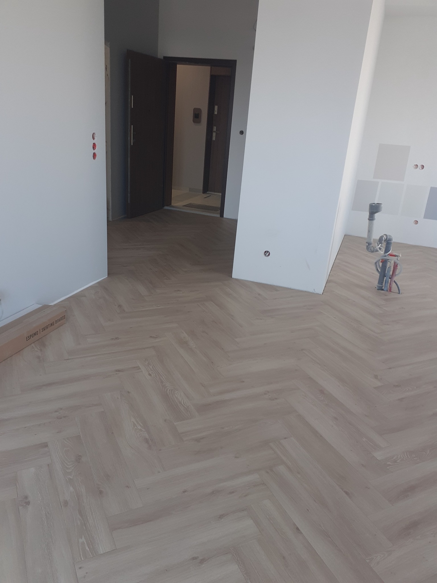 Jasne panele ułożone w jodełkę francuską w nowym mieszkaniu, widoczny karton z listwami przypodłogowymi i narzędzia w tle.