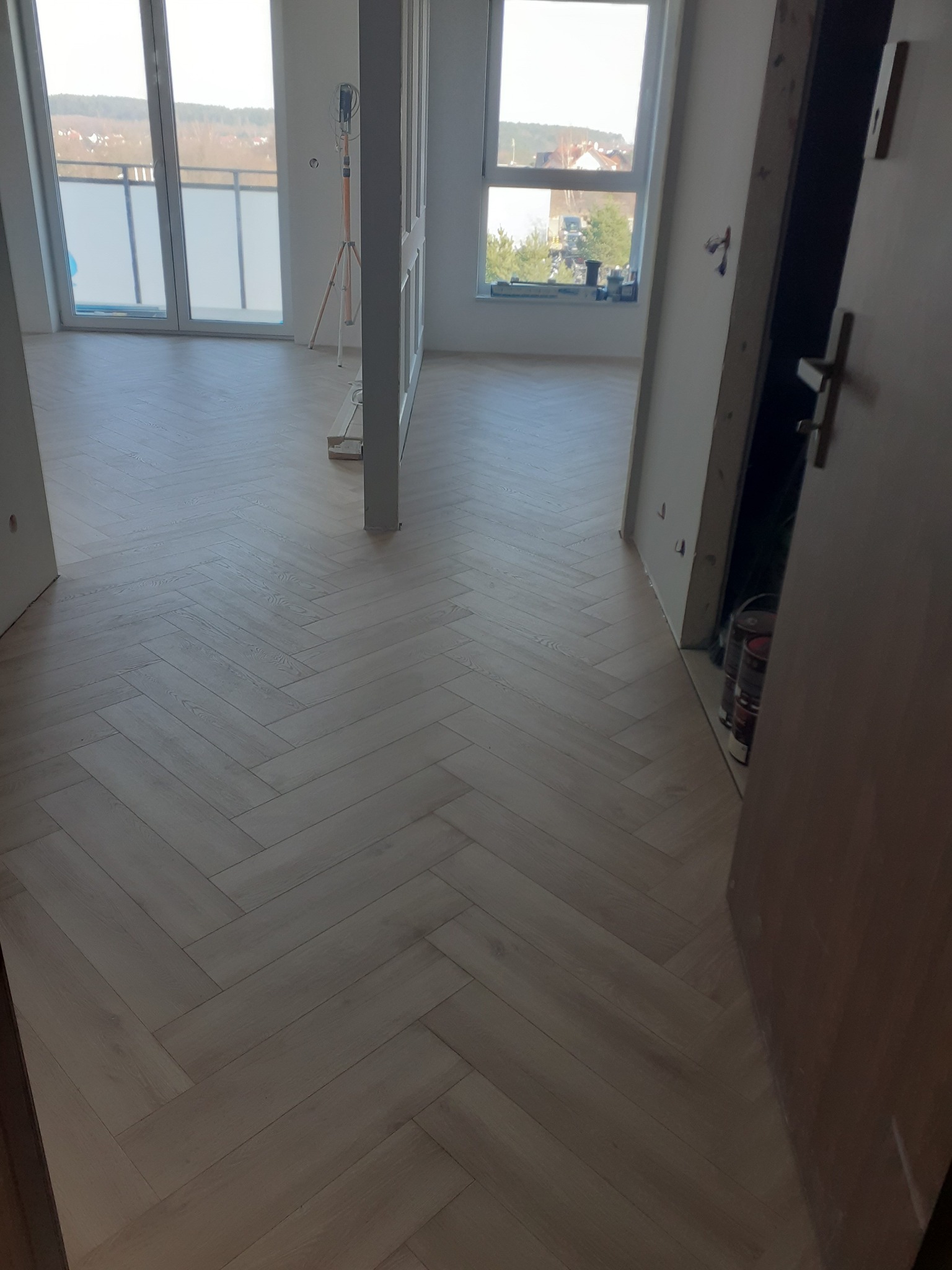 Jasne panele ułożone we wzór jodełki francuskiej w nowym, jasnym mieszkaniu, widok na okno i drzwi w trakcie montażu.