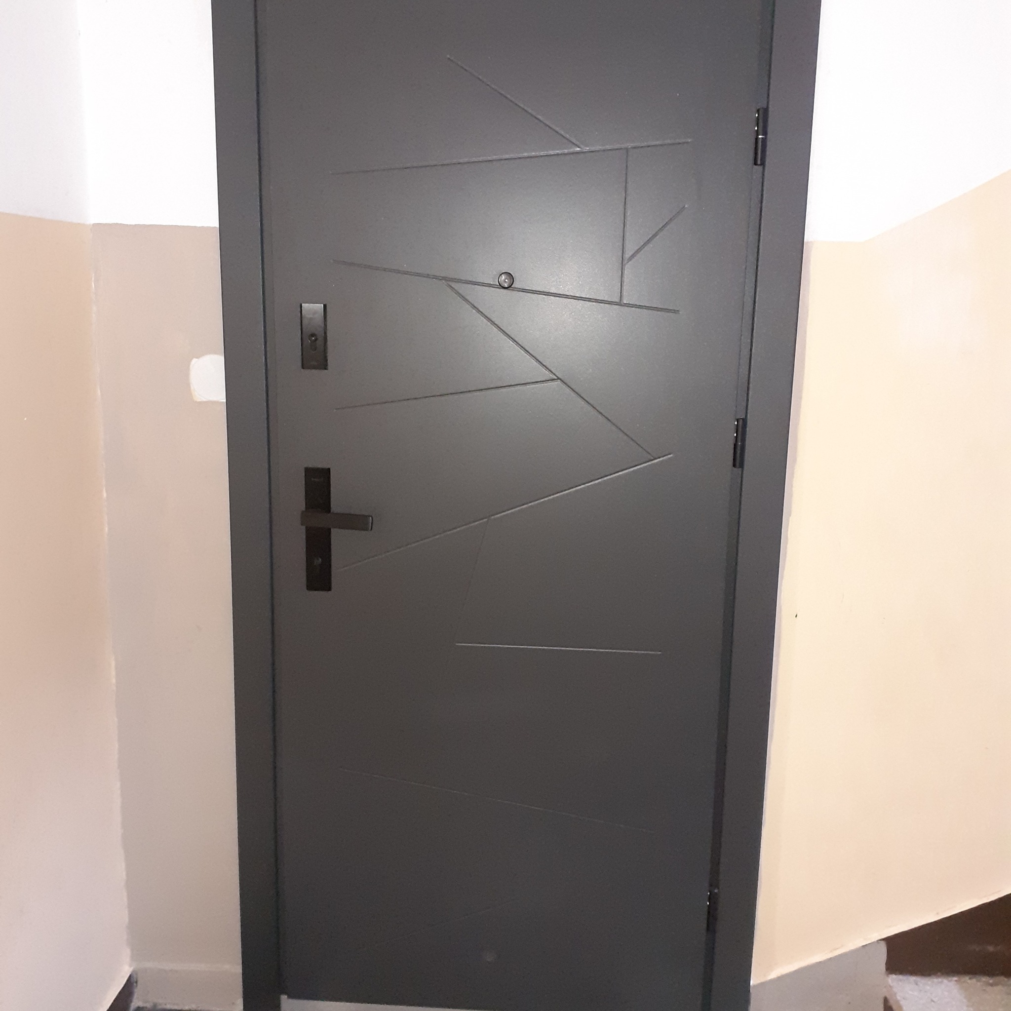 Grafitowe drzwi wejściowe z geometrycznym wzorem, czarną klamką i wizjerem, zamontowane w klatce schodowej z beżowymi ścianami.