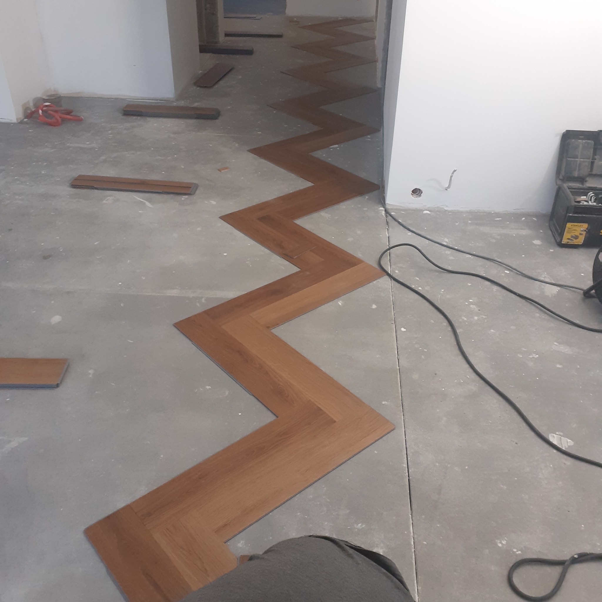 Fragment układania paneli podłogowych w jodełkę na surowym, betonowym podłożu; widoczne narzędzia w skrzynce i luźne panele.