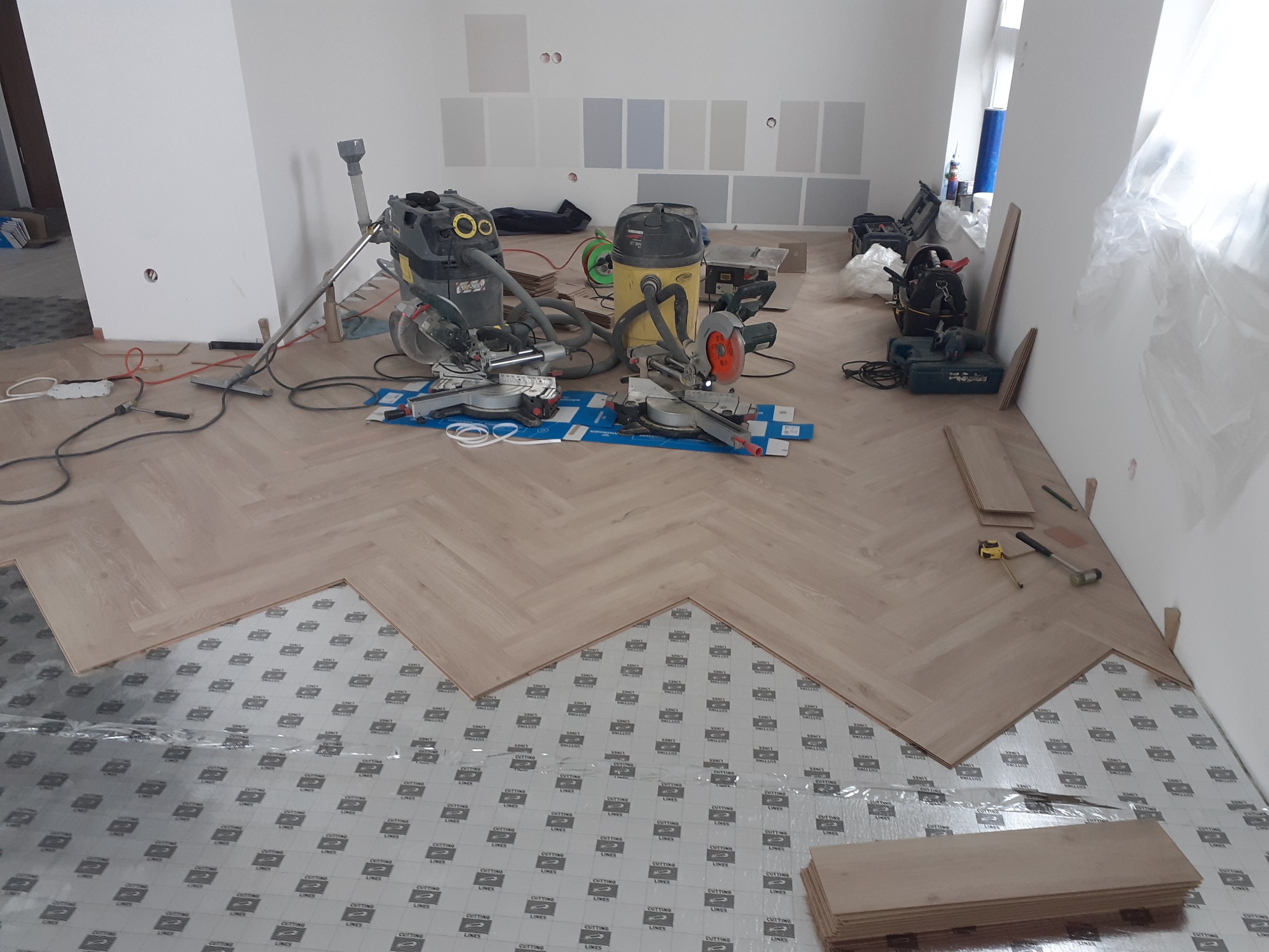 Fragment pokoju w trakcie układania parkietu w jodełkę; widoczne narzędzia, odkurzacze przemysłowe i podkład pod panele.