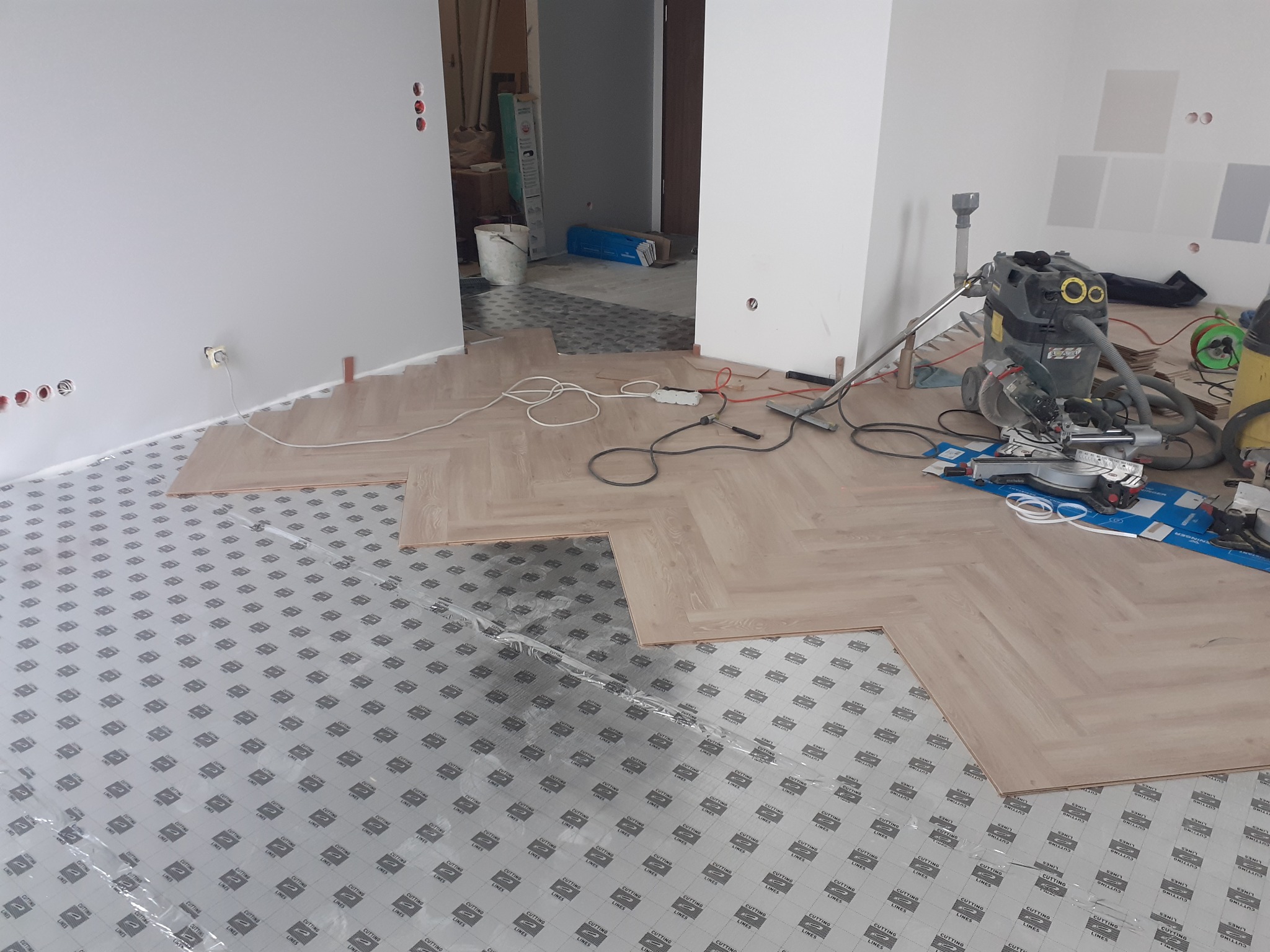 Układanie jasnych paneli podłogowych w jodełkę na podkładzie z folii ochronnej, widoczny obszar roboczy z narzędziami, odkurzaczem przemysłowym i niedokończoną podłogą w tle.