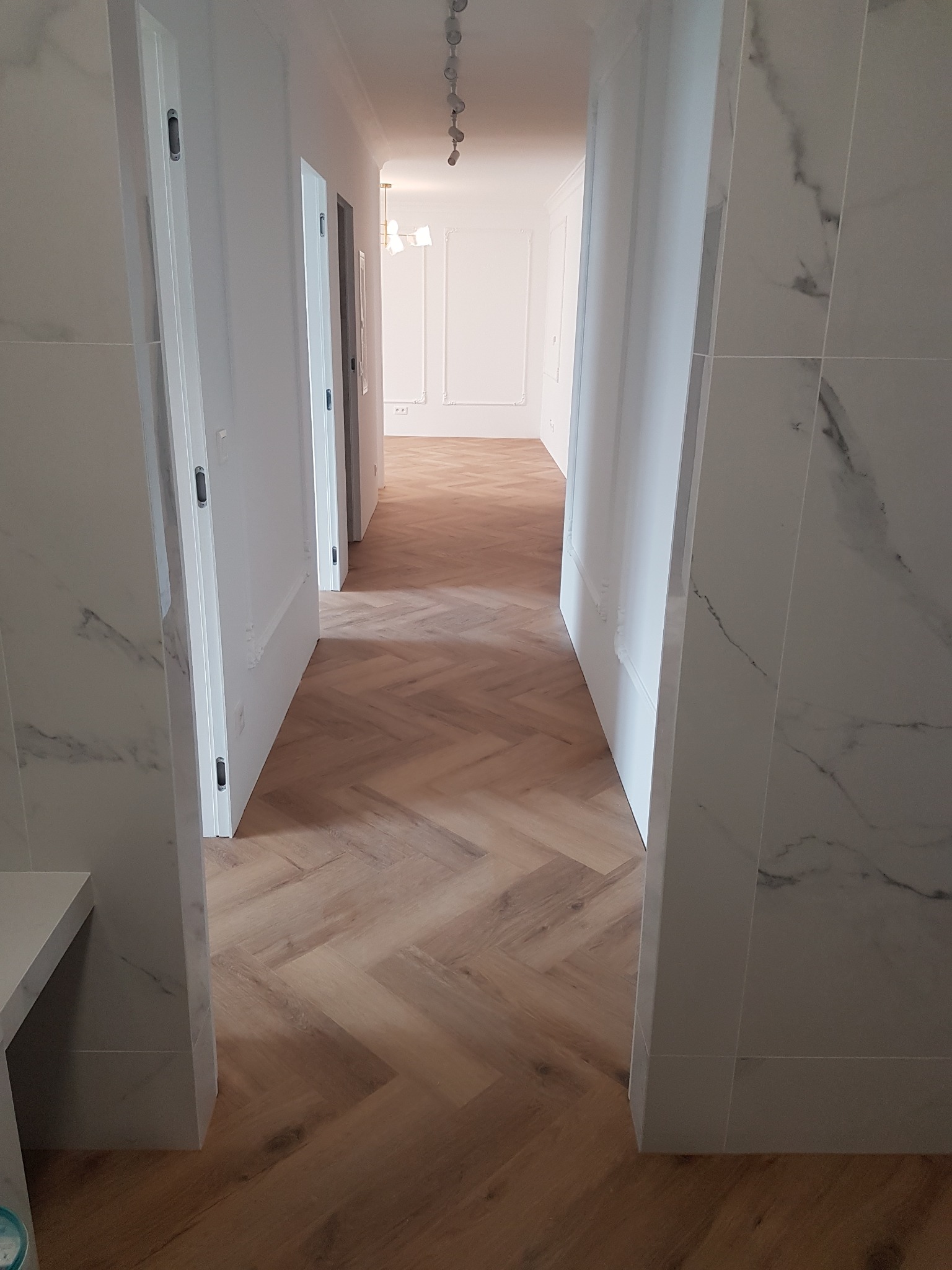 Korytarz z parkietem ułożonym w jodełkę, widok przez otwarte białe drzwi z marmurowym wykończeniem