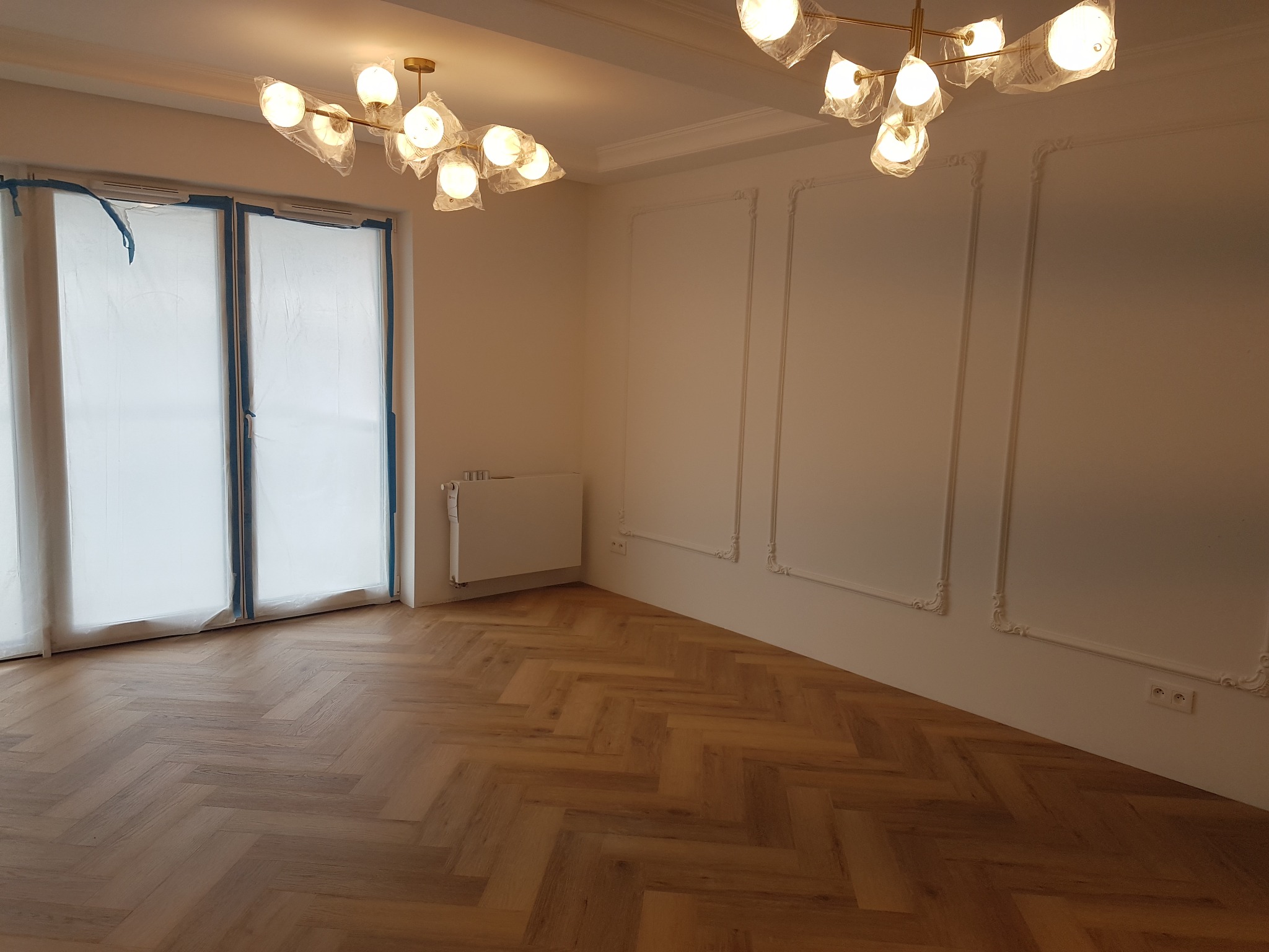 Ułożona podłoga z paneli w jodełkę, okna i drzwi zabezpieczone folią malarską, nowoczesne lampy na suficie, dekoracyjne sztukaterie na ścianach.