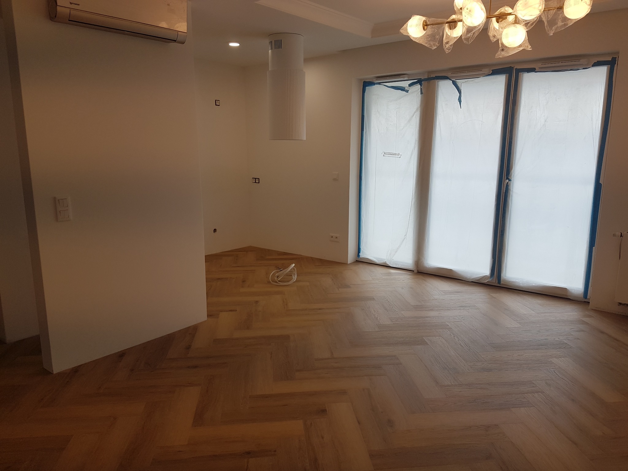 Ukończone układanie parkietu w jodełkę w jasnym wnętrzu, okna zabezpieczone folią malarską, widoczne wystające kable elektryczne.