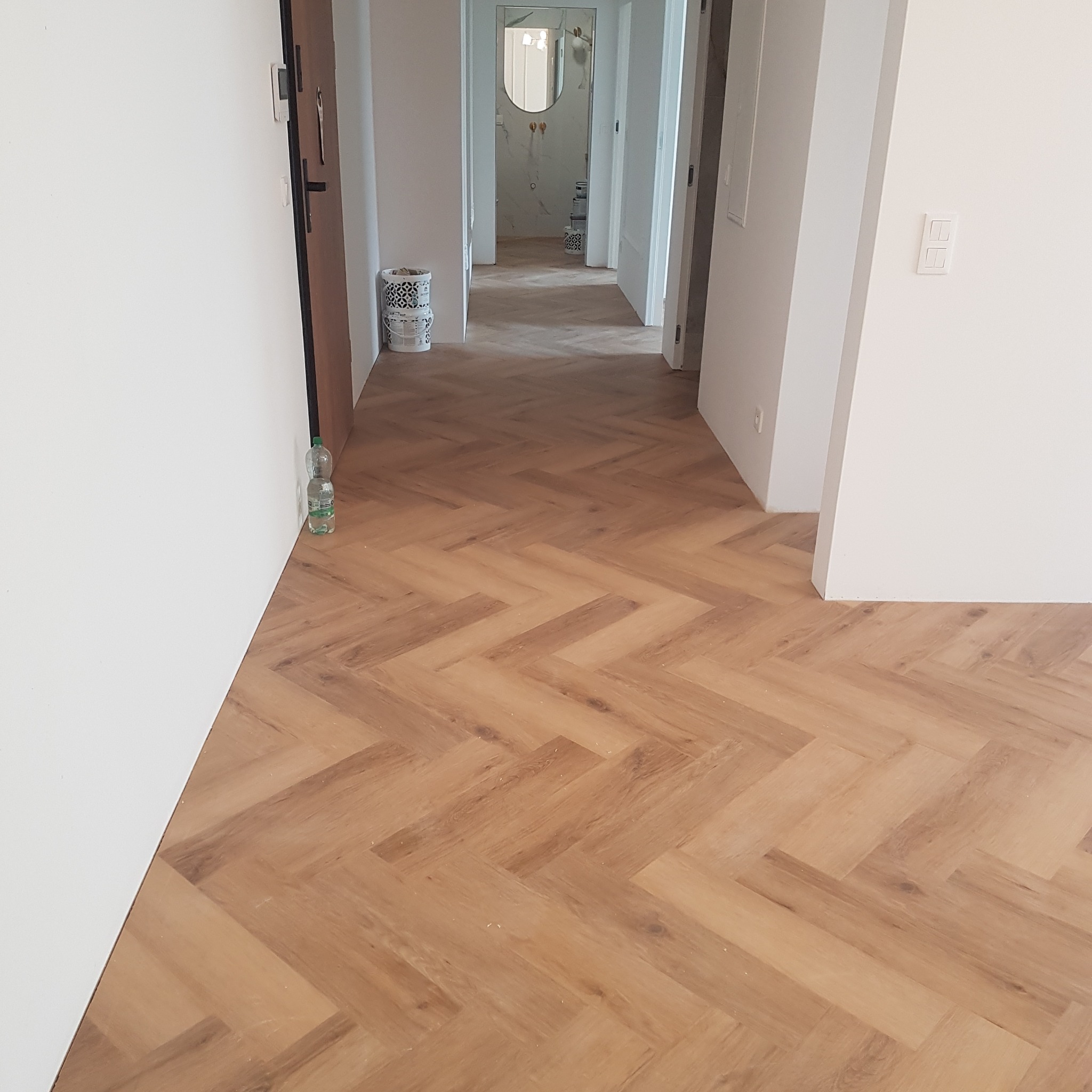 Ułożona podłoga z paneli w jodełkę, widok na korytarz z białymi ścianami i butelką wody przy ścianie.