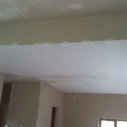 Wnętrze w stanie deweloperskim po nałożeniu jasnych tynków na ścianach i suficie, widoczne schody w tle.